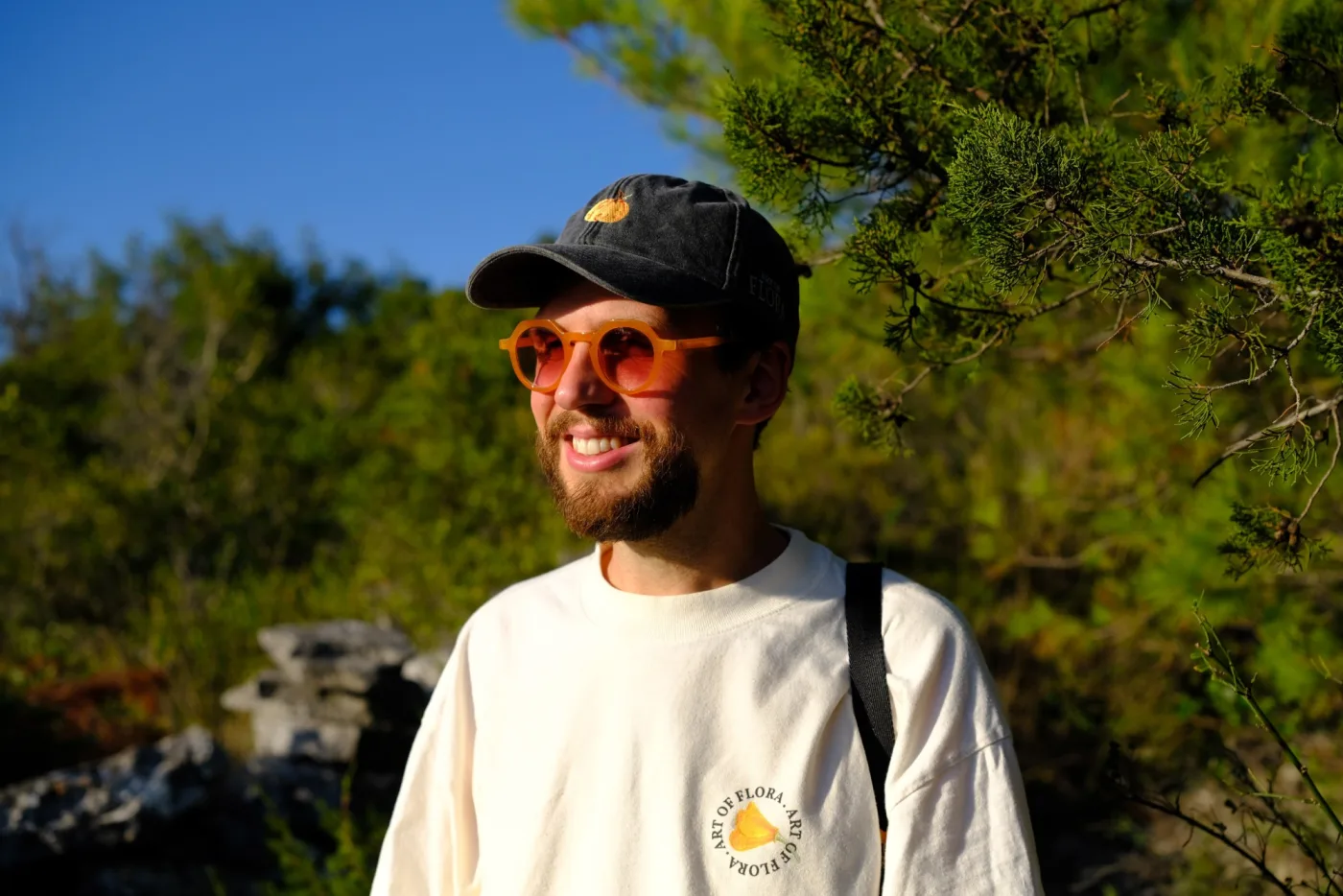 A smiling man with a beard, wearing orange sunglasses, a dark cap with an embroidered orange flower and a cream-coloured Art of Flora shirt, standing in front of green trees
